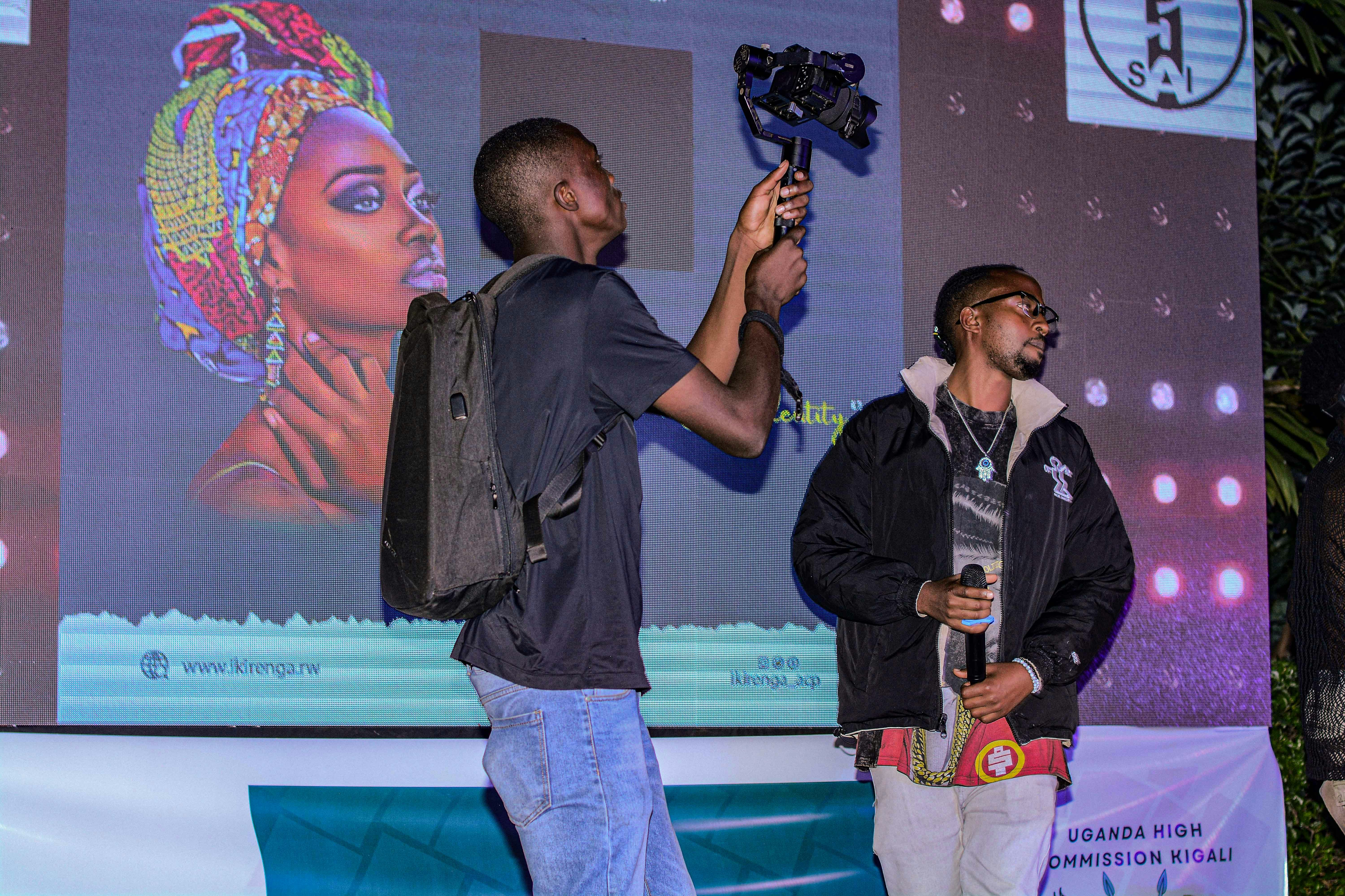 A person wearing a backpack is holding a camera-mounted stabilizer, standing beside another individual holding a microphone. They are positioned in front of a large screen displaying vibrant artwork of a woman with a colorful headscarf. The scene appears to be taking place at an event, possibly a presentation or performance.