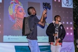 A person wearing a backpack is holding a camera-mounted stabilizer, standing beside another individual holding a microphone. They are positioned in front of a large screen displaying vibrant artwork of a woman with a colorful headscarf. The scene appears to be taking place at an event, possibly a presentation or performance.