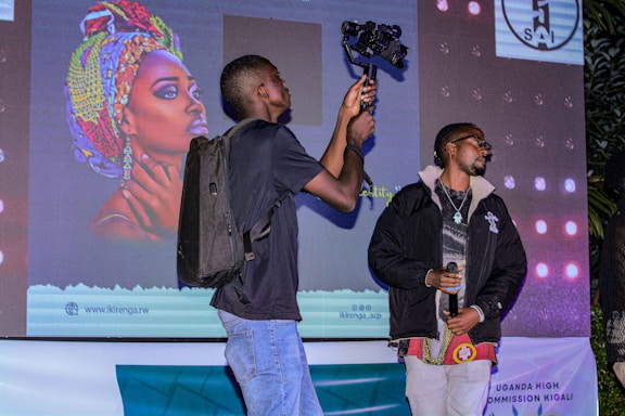 A person wearing a backpack is holding a camera-mounted stabilizer, standing beside another individual holding a microphone. They are positioned in front of a large screen displaying vibrant artwork of a woman with a colorful headscarf. The scene appears to be taking place at an event, possibly a presentation or performance.