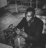 A person is sitting indoors, adjusting a professional camera mounted on a tripod. The setting appears to be a casual space with tiled flooring and a metal chair nearby. The image is in black and white, providing a timeless feel.
