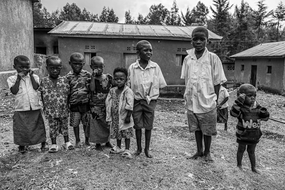 A group of young children stands together outdoors in front of simple, rural buildings. They are dressed in casual clothes, and their expressions vary from curious to neutral. The background features several modest structures with tin roofs, set against a backdrop of trees.