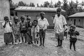 A group of young children stands together outdoors in front of simple, rural buildings. They are dressed in casual clothes, and their expressions vary from curious to neutral. The background features several modest structures with tin roofs, set against a backdrop of trees.