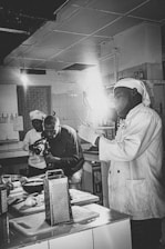 Two people are in a kitchen setting, with one person wearing a chef's uniform holding a utensil, and the other person is filming with a camera. The area is equipped with various kitchen tools and appliances, including a grater, chopping board, and bowls.