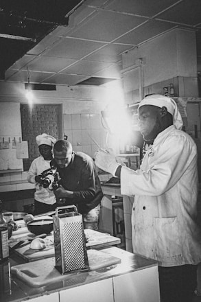 Two people are in a kitchen setting, with one person wearing a chef's uniform holding a utensil, and the other person is filming with a camera. The area is equipped with various kitchen tools and appliances, including a grater, chopping board, and bowls.
