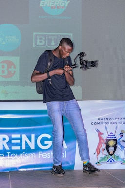 A person holding a camera rig stands on a stage with a backdrop displaying various logos, including one for a Uganda tourism festival. The person wears a black shirt, jeans, and black shoes. There is a banner with the Uganda High Commission logo and text partially visible.