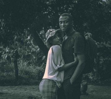 A couple standing closely together in an outdoor setting. The woman wears a hat and plaid pants, while the man has a backpack slung over one shoulder. The background features dense foliage and appears to be a wooded area, with subdued lighting suggesting an intimate or serene atmosphere.