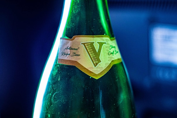 A close-up view of a dark green glass bottle with a label featuring the letter 'V' in a vintage style. The label has text indicating 'Artisanal Recipe Beer' and is set against a blurred blue and black background.
