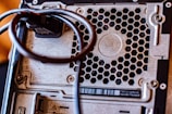 Close-up of a computer's back panel featuring a metal honeycomb grill, screws, and a cable looped around a connector. The metallic surface shows some signs of wear.