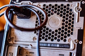 Close-up of a computer's back panel featuring a metal honeycomb grill, screws, and a cable looped around a connector. The metallic surface shows some signs of wear.