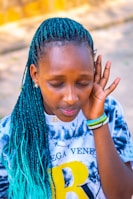 A person with closed eyes and teal braided hair wears a tie-dye shirt with a large yellow letter 'B' on it. They have bracelets on their wrist and appear to be listening intently or focusing on a sound.