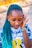 A person with closed eyes and teal braided hair wears a tie-dye shirt with a large yellow letter 'B' on it. They have bracelets on their wrist and appear to be listening intently or focusing on a sound.