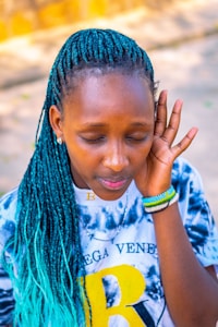 A person with closed eyes and teal braided hair wears a tie-dye shirt with a large yellow letter 'B' on it. They have bracelets on their wrist and appear to be listening intently or focusing on a sound.