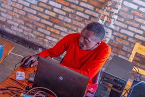 A person wearing a bright red shirt is seated at a wooden table, using a laptop. The setting appears to be indoors with a background of textured brick walls. Various electronic devices, including a computer tower and some cables, are scattered on the table.