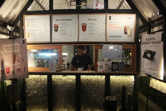 A coffee and tea stall with various promotional posters and menus displayed. There's a man standing behind the counter inside a wooden-framed cubicle. The interior is warmly lit, and modern design elements are present.
