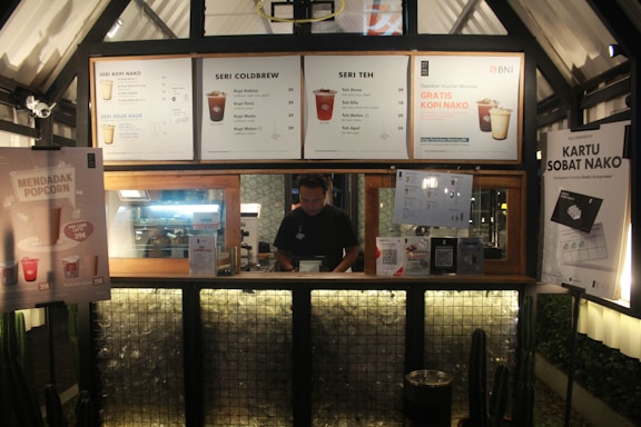 A coffee and tea stall with various promotional posters and menus displayed. There's a man standing behind the counter inside a wooden-framed cubicle. The interior is warmly lit, and modern design elements are present.