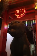 A stone statue of a lion is positioned in an outdoor setting with an illuminated background featuring a red neon sign shaped like a ‘W’. Colorful hanging lights add vibrancy to the scene, and overhead wooden beams suggest a structural feature like a pavilion or gateway.