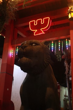 A stone statue of a lion is positioned in an outdoor setting with an illuminated background featuring a red neon sign shaped like a ‘W’. Colorful hanging lights add vibrancy to the scene, and overhead wooden beams suggest a structural feature like a pavilion or gateway.