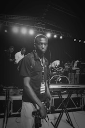 A person with a media badge is standing near a stage holding two cameras. Behind them, there are musical instruments like a Yamaha keyboard and a drum set. Several other people are visible in the background, engaged in various activities.