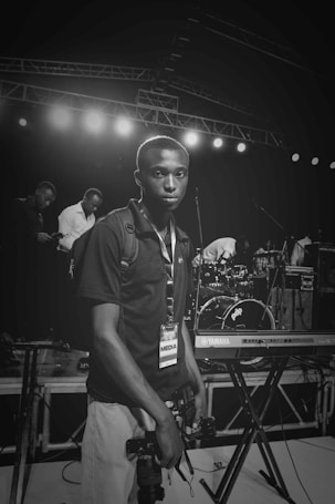 A person with a media badge is standing near a stage holding two cameras. Behind them, there are musical instruments like a Yamaha keyboard and a drum set. Several other people are visible in the background, engaged in various activities.