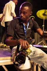 A young man holding a Canon camera sits with a focused expression. He is wearing a black shirt, light blue jeans, and a blue lanyard around his neck. Behind him, a drum set and other people are visible, suggesting a live event or performance setting.
