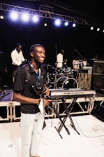 A person smiles on stage holding a camera and a camera rig, surrounded by musical instruments such as a keyboard, drum set, and amplifiers. Several other individuals are positioned behind him, seemingly setting up or preparing for a performance. Bright stage lights illuminate the scene from above.