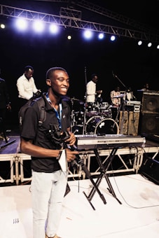 A person smiles on stage holding a camera and a camera rig, surrounded by musical instruments such as a keyboard, drum set, and amplifiers. Several other individuals are positioned behind him, seemingly setting up or preparing for a performance. Bright stage lights illuminate the scene from above.