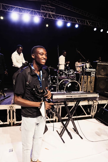 A person smiles on stage holding a camera and a camera rig, surrounded by musical instruments such as a keyboard, drum set, and amplifiers. Several other individuals are positioned behind him, seemingly setting up or preparing for a performance. Bright stage lights illuminate the scene from above.