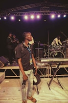 A man with a media badge stands in the foreground holding camera equipment on a stage set with musical instruments. Behind him are a keyboard and drum set, while additional people in the background appear to be setting up or preparing for a performance. Stage lighting and trusses are visible overhead.