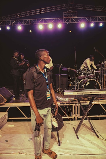 A man with a media badge stands in the foreground holding camera equipment on a stage set with musical instruments. Behind him are a keyboard and drum set, while additional people in the background appear to be setting up or preparing for a performance. Stage lighting and trusses are visible overhead.