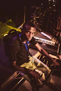A person sits on stage, holding a camera, surrounded by musical instruments like a keyboard and drum set. The lighting is dim, with a focus on the individual, suggesting a pre-performance or backstage setting.