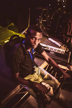 A person sits on stage, holding a camera, surrounded by musical instruments like a keyboard and drum set. The lighting is dim, with a focus on the individual, suggesting a pre-performance or backstage setting.
