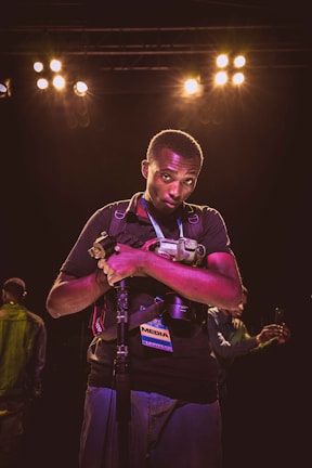 A person holding a camera and resting their arms on a monopod is standing under bright stage lights, with a media pass hanging around their neck. The background shows another person on the left taking a photo with a smartphone.