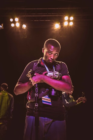 A person holding a camera and resting their arms on a monopod is standing under bright stage lights, with a media pass hanging around their neck. The background shows another person on the left taking a photo with a smartphone.