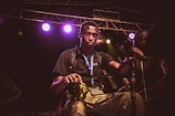 A young man wearing a black shirt and a blue lanyard with a media badge is holding a yellow DSLR camera. He is surrounded by camera equipment and lights, indicating a professional environment, possibly a concert or event. The background shows stage lighting with multiple colored lights and scaffolding.