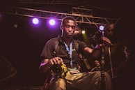 A young man wearing a black shirt and a blue lanyard with a media badge is holding a yellow DSLR camera. He is surrounded by camera equipment and lights, indicating a professional environment, possibly a concert or event. The background shows stage lighting with multiple colored lights and scaffolding.