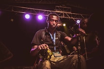A young man wearing a black shirt and a blue lanyard with a media badge is holding a yellow DSLR camera. He is surrounded by camera equipment and lights, indicating a professional environment, possibly a concert or event. The background shows stage lighting with multiple colored lights and scaffolding.