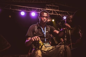 A young man wearing a black shirt and a blue lanyard with a media badge is holding a yellow DSLR camera. He is surrounded by camera equipment and lights, indicating a professional environment, possibly a concert or event. The background shows stage lighting with multiple colored lights and scaffolding.