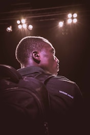 A person is seen from a side profile under dramatic stage lighting. They are wearing a dark backpack and a dark jacket with 'Under Armour' written on it. Bright stage lights illuminate their face and create a glowing outline.