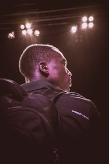 A person is seen from a side profile under dramatic stage lighting. They are wearing a dark backpack and a dark jacket with 'Under Armour' written on it. Bright stage lights illuminate their face and create a glowing outline.