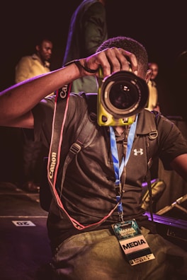 A person is taking a photograph using a DSLR camera with a yellow body, prominently facing the camera. The person is wearing a black shirt with a recognizable sports logo and a blue lanyard that holds a media pass around their neck. A red camera strap is visible and the background shows several other people in dim lighting.