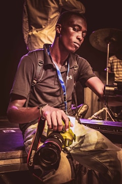 A person is sitting on the edge of a stage holding a Canon camera. The individual is wearing a dark polo shirt, a backpack, and has a blue lanyard around the neck. Musical instruments, including a drum set, are visible in the background.