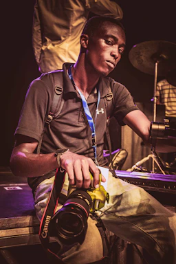 A person is sitting on the edge of a stage holding a Canon camera. The individual is wearing a dark polo shirt, a backpack, and has a blue lanyard around the neck. Musical instruments, including a drum set, are visible in the background.