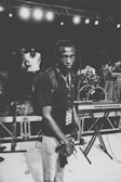 A black and white photograph featuring a person with a media pass around their neck and holding a camera. The background displays a music stage setup with a keyboard and drums. There are other individuals, likely musicians, engaged in conversation.