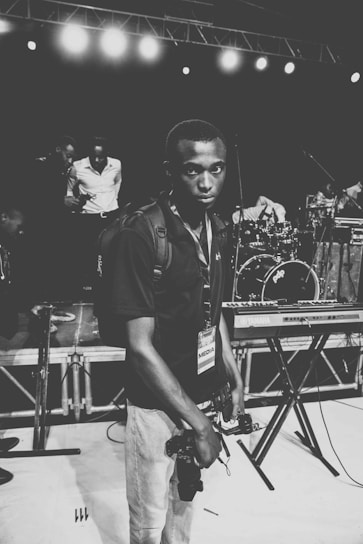 A black and white photograph featuring a person with a media pass around their neck and holding a camera. The background displays a music stage setup with a keyboard and drums. There are other individuals, likely musicians, engaged in conversation.