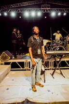 Several people are on a stage, engaged in different activities. One person stands at the forefront holding a camera, wearing a media pass around their neck. In the background, some individuals are adjusting musical equipment, including a drum set and keyboard. The stage is well-lit with multiple overhead lights and there is a metal truss setup above.