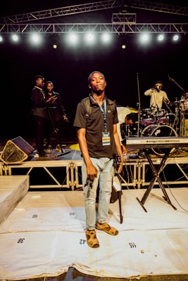 Several people are on a stage, engaged in different activities. One person stands at the forefront holding a camera, wearing a media pass around their neck. In the background, some individuals are adjusting musical equipment, including a drum set and keyboard. The stage is well-lit with multiple overhead lights and there is a metal truss setup above.