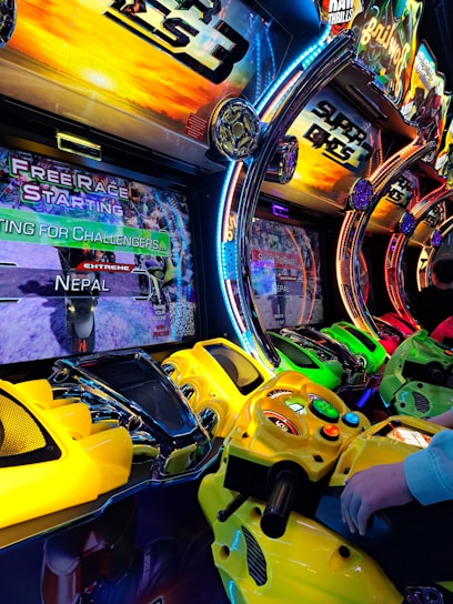 A row of colorful arcade motorcycle racing game machines with bright LED lights and vibrant yellow, green, and red controllers. The display screens show racing graphics and text such as 'Free Race Starting' and 'Extreme Nepal.' The setting has an energetic atmosphere with vivid game themes and a player holding onto one of the game's handlebars.