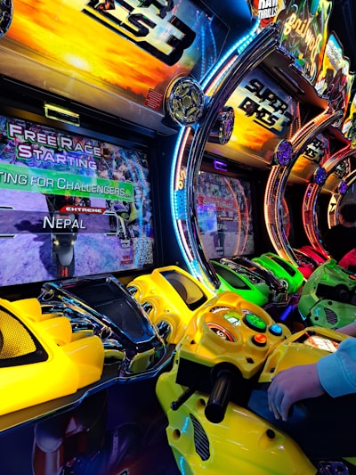 A row of colorful arcade motorcycle racing game machines with bright LED lights and vibrant yellow, green, and red controllers. The display screens show racing graphics and text such as 'Free Race Starting' and 'Extreme Nepal.' The setting has an energetic atmosphere with vivid game themes and a player holding onto one of the game's handlebars.
