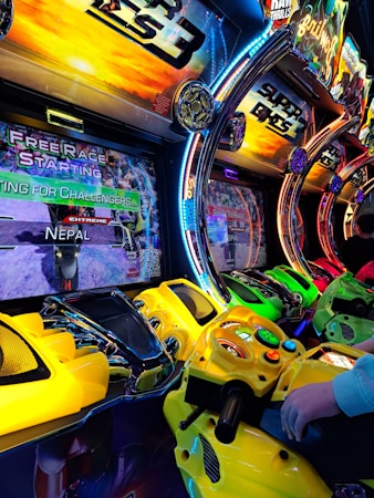 A row of colorful arcade motorcycle racing game machines with bright LED lights and vibrant yellow, green, and red controllers. The display screens show racing graphics and text such as 'Free Race Starting' and 'Extreme Nepal.' The setting has an energetic atmosphere with vivid game themes and a player holding onto one of the game's handlebars.