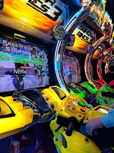 A row of colorful arcade motorcycle racing game machines with bright LED lights and vibrant yellow, green, and red controllers. The display screens show racing graphics and text such as 'Free Race Starting' and 'Extreme Nepal.' The setting has an energetic atmosphere with vivid game themes and a player holding onto one of the game's handlebars.
